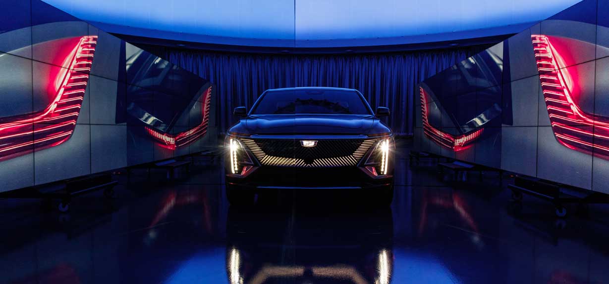 Front end of 2023 Cadillac LYRIQ EV with headlights on and mirror reflection from floor of stage